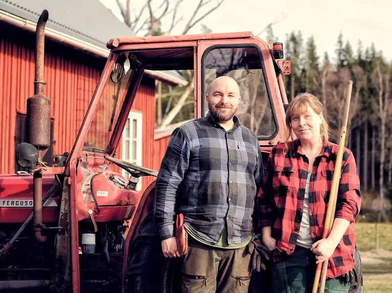 Två personer som står bredvid varandra framför en traktor, iförda kläder lämpliga för trädgårdsarbete. Personerna är Fredrik och Johanna, som driver Fagralund Trädgård.
